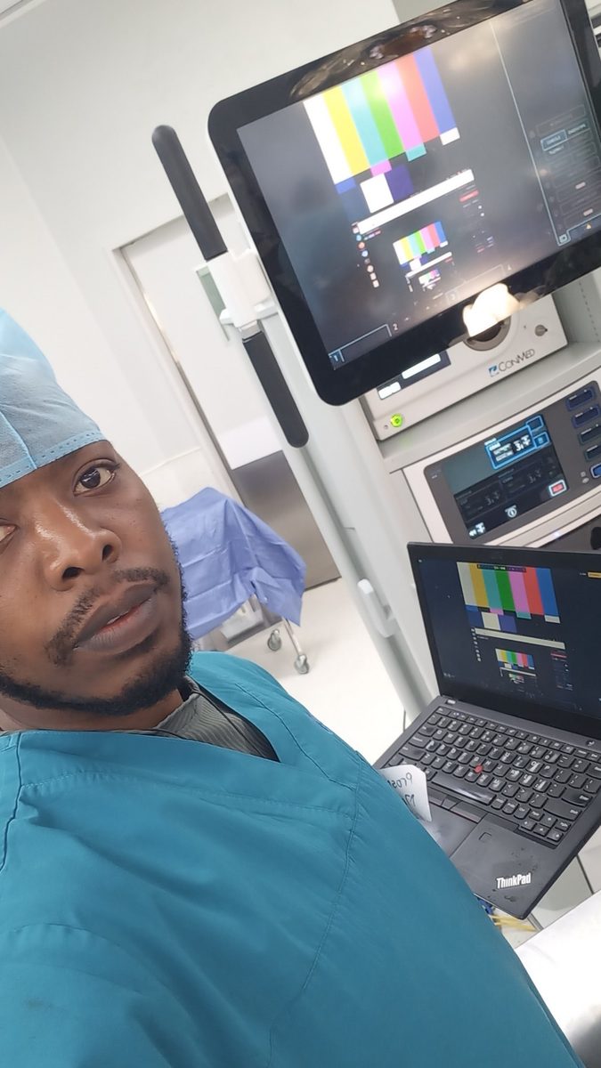 Prosper in scrubs beside an endoscopy tower showing colour bars